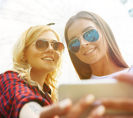 Zwei Frauen mit Sonnenbrillen im Sonnenschein machen ein Selfie mit einem weißen Smartphone
