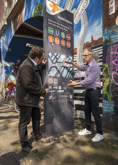 Minister Oliver Krischer und Dr. Norbert Reinkober schauen auf die Stele der Mobilstation am Bahnhof Köln-Ehrenfeld.