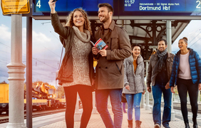 Ein junges Paar steht an einem Bahnsteig und macht ein Selfie. Im Hintergrund kommt eine weitere Gruppe Personen auf sie zugelaufen.