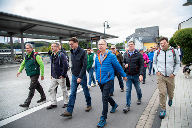 Verkehrsminister Hendrik Wüst, Aktionspate Manuel Andrack wandern mit einer Gruppe Menschen über das Bahnhofsgelände Overath.