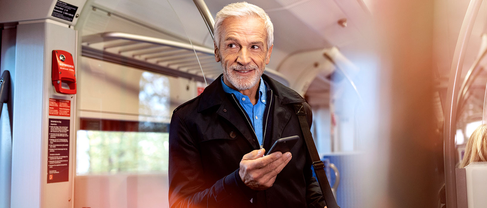 Ein gut gekleideter Senior steht in einer Bahn und schaut auf sein Handy.