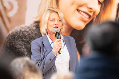 Birgit Strecker vom KCM NRW steht vor einem Plakat der App und spricht in ein Mikrofon.