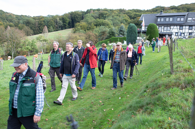 Eine Wandergruppe mit Manuel Andrack läuft auf einem Wiesenweg, im Hintergrund ist ein Gasthaus zu sehen.
