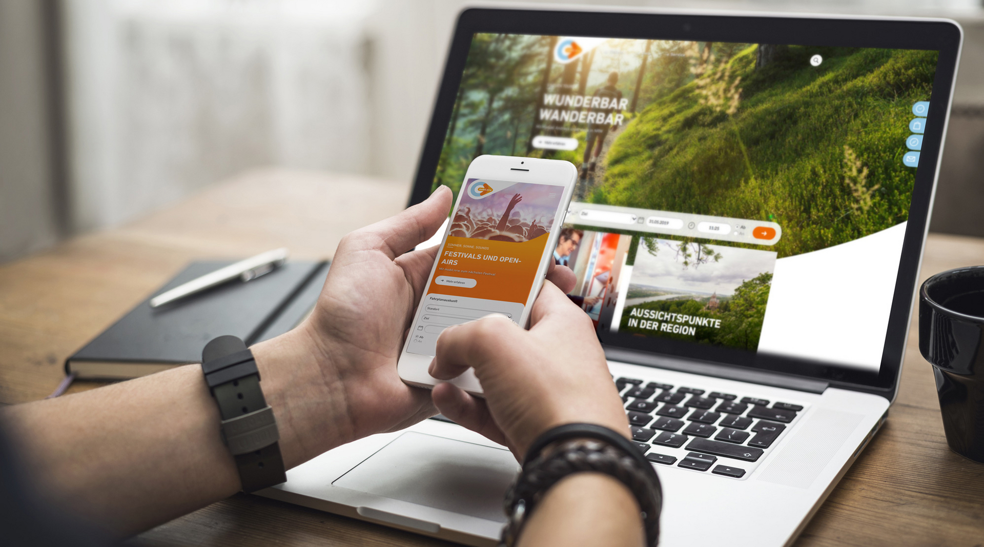 Zwei Hände halten ein weißes Smartphone vor einem Laptop. Auf dem Handy ist die mobil.nrw App zu sehen mit einer Aktion zur Festivalsaison. Auf dem Laptop Bildschirm ist die mobil.nrw Desktop Seite zu sehen mit einem Wander-Spezial.