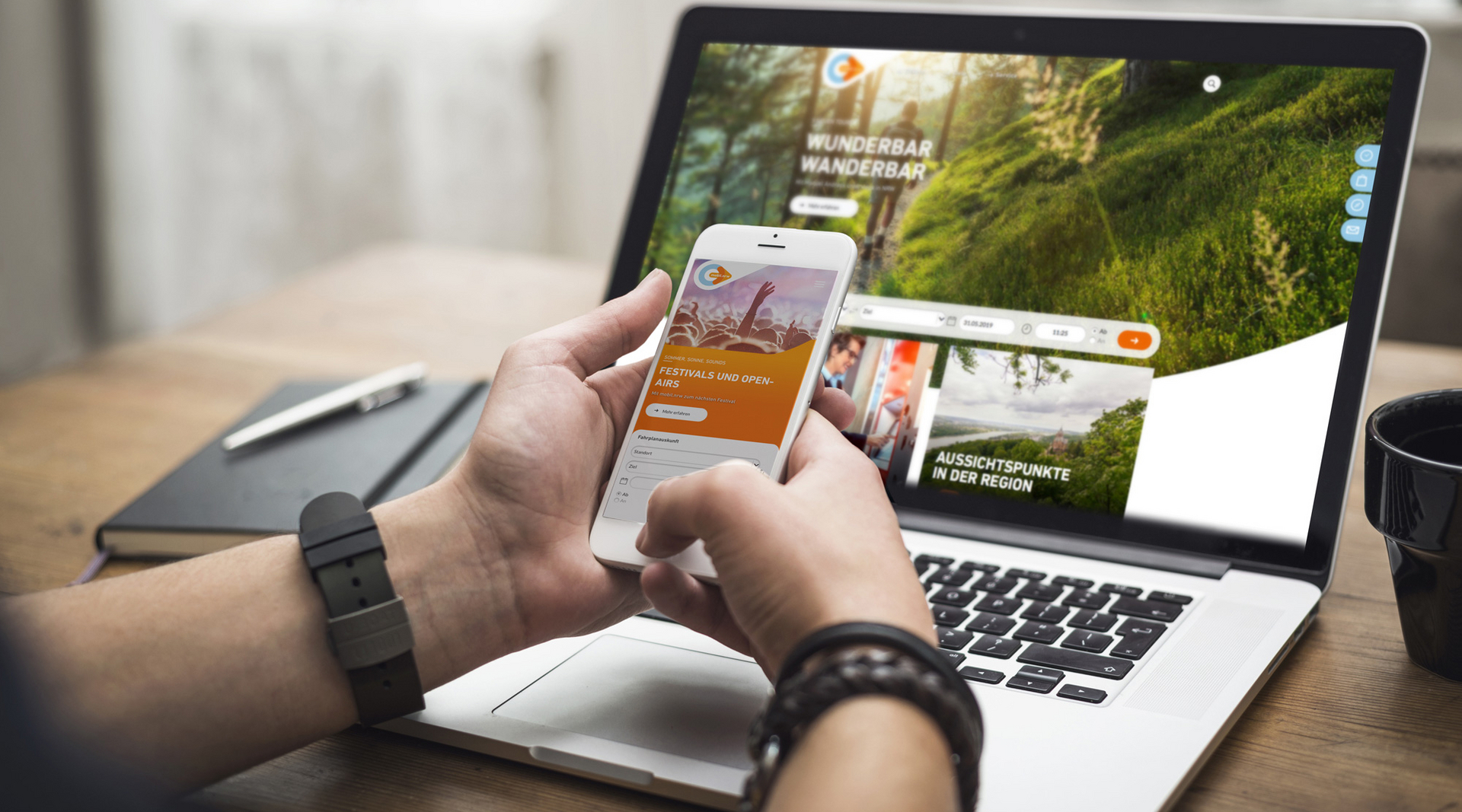Zwei Hände halten ein weißes Smartphone vor einem Laptop. Auf dem Handy ist die mobil.nrw App zu sehen mit einer Aktion zur Festivalsaison. Auf dem Laptop Bildschirm ist die mobil.nrw Desktop Seite zu sehen mit einem Wander-Spezial.