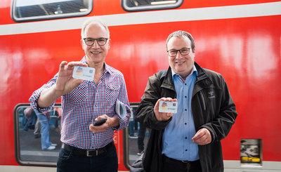 Dr. Norbert Reinkober und Minister Oliver Krischer stehen vor einem roten Regionalzug und halten Deutschlandtickets in der Hand.
