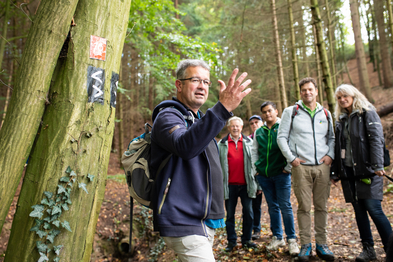 Manuel Andrack steht vor einer Menschengruppe im Wald bei Overath und erklärt etwas.
