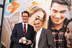 NRW-Verkehrsminister Hendrik Wüst steht neben einem Mann vor einem Plakat der App und hält ein Handy in der Hand, auf dem die App geöffnet ist.