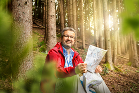 Manuel Andrack sitzt mit einer Wanderkarte in einem Wald, durch dessen Bäume Sonnenstrahlen scheinen.