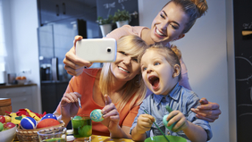 Drei Frauen unterschiedlichen Alters sitzen am Küchentisch und bemalen Ostereier, während sie über ein Handy ein Selfie machen.