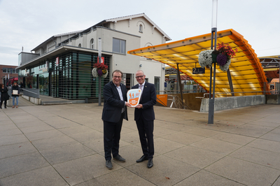 Oliver Krischer, Minister für Umwelt, Naturschutz und Verkehr NRW (links) und Oliver Kellner, Bürgermeister der Stadt Emsdetten, stehen mit der Wanderbahnhofsplakette vor dem Bahnhofsgebäude Emsdetten.