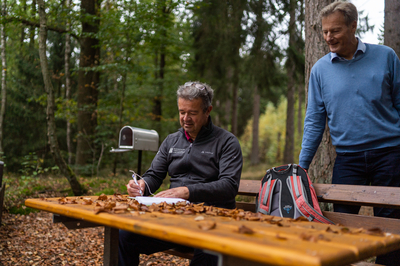 Manuel Andrack sitzt an einem Holztisch und schreibt in ein Wanderbuch, ein anderer Mann steht hinter ihm und schaut ihm zu.