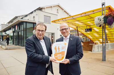 Oliver Krischer, Minister für Umwelt, Naturschutz und Verkehr NRW (links) und Oliver Kellner, Bürgermeister der Stadt Emsdetten, stehen mit der Wanderbahnhofsplakette vor dem Bahnhofsgebäude Emsdetten.